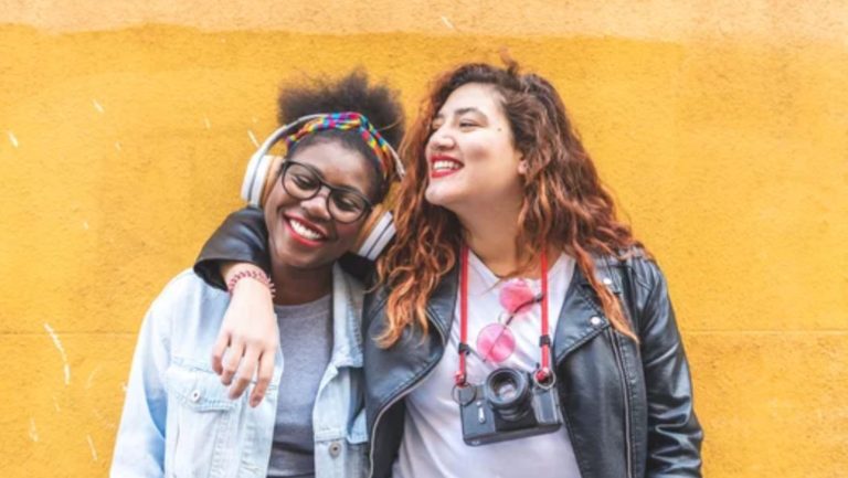 Two smiling young women standing together against a yellow wall, one wearing over-ear headphones and the other carrying a camera.