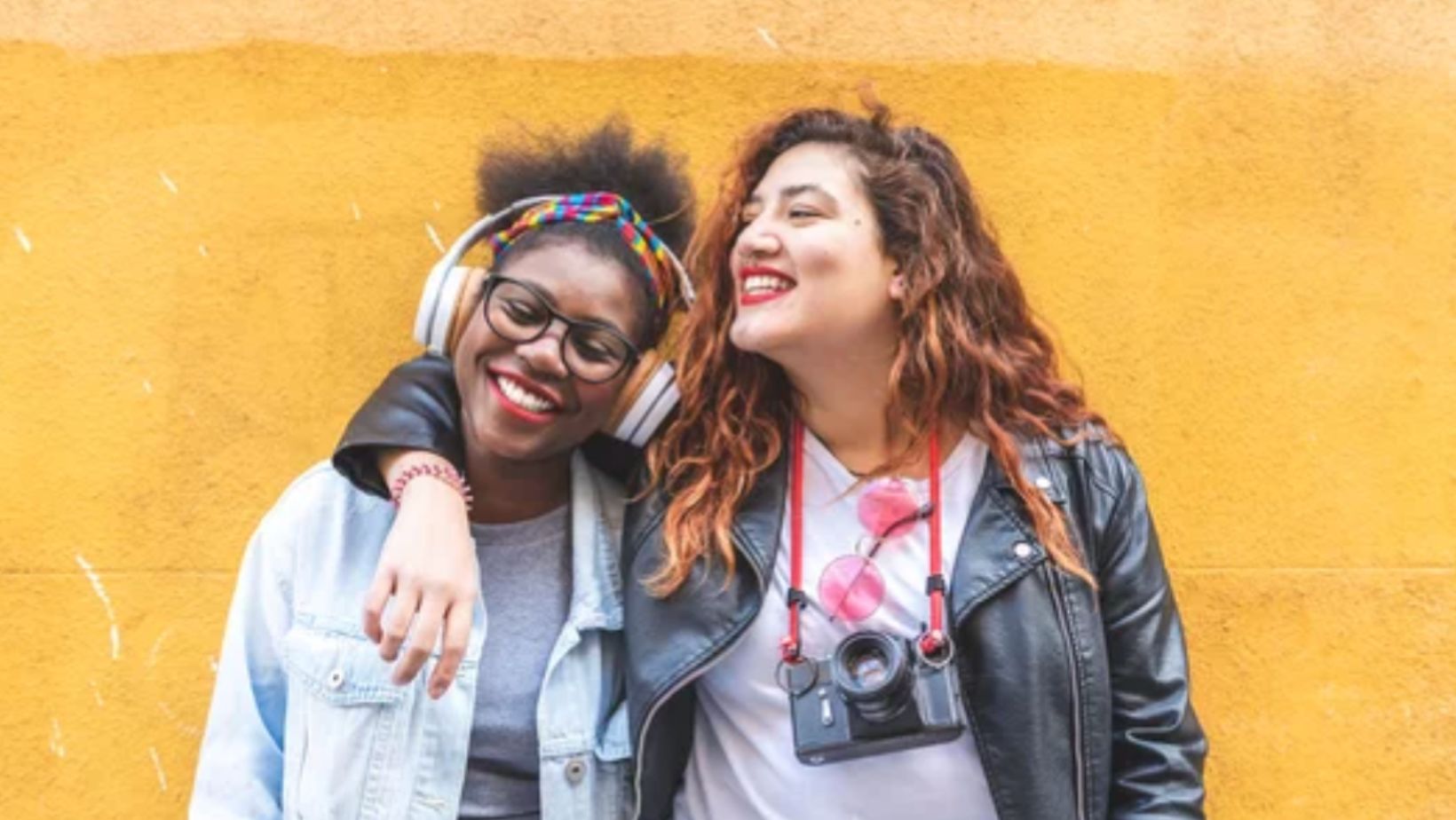 Two smiling young women standing together against a yellow wall, one wearing over-ear headphones and the other carrying a camera.